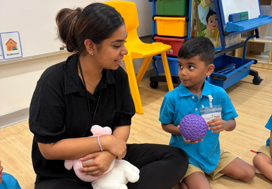 Woman seated on floor talks to young boy holding a purple ball in a classroom with yellow chairs.